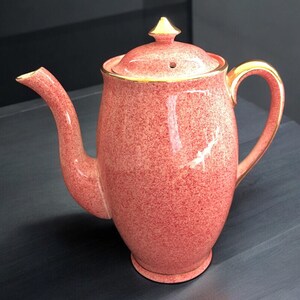 May include: A vintage-style, speckled pink ceramic teapot with a lid and gold-coloured handle and trim. The teapot has a long spout and a rounded body. The lid is topped with a gold finial. A classic piece for serving tea.