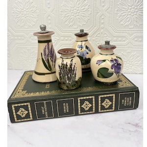 May include: Three ceramic shakers with metal tops, each featuring hand-painted floral designs including lavender and violets. The shakers are arranged on a dark green hardcover book titled Great Expectations by Charles Dickens with gold lettering.