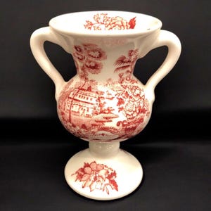 May include: A white ceramic vase with two handles and a red floral pattern. The vase has a pedestal base and features a red and white floral design.