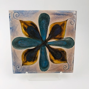May include: Square ceramic tile featuring a floral design. The flower has blue and yellow petals, with a blue centre. The background is a light beige with a blue border and decorative swirls in each corner.