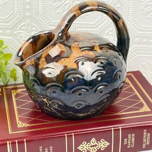 May include: A round ceramic pitcher with a single handle, featuring a glossy glaze in shades of deep blue and warm brown. The lower portion is decorated with a repeating embossed wave pattern, while the upper section remains smooth.