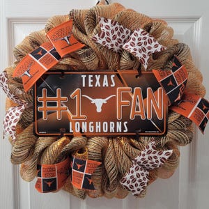 May include: A decorative wreath featuring a metallic mesh base, adorned with orange and white ribbons, and football-themed accents. The centerpiece is a license plate that reads "#1 FAN LONGHORNS" with the Texas Longhorns logo.