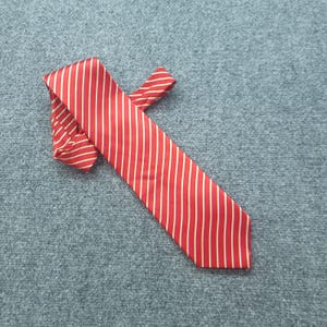 Puede incluir: Una corbata de color rojo vibrante con finas rayas diagonales blancas que recorren toda la longitud de la tela. La corbata está doblada sobre sí misma, mostrando una textura suave y satinada con un extremo puntiagudo en la parte inferior.