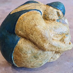 Könnte beinhalten: Eine Keramikskulptur einer Person in einem blauen Badeanzug, die sich in fötaler Position zusammengerollt hat. Die Skulptur ist in Gelb- und Blautönen bemalt.