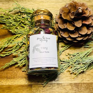 May include: A glass jar filled with dried herbs and berries labelled "Honey Creek Witchery Yule Ritual Herbs". The jar is sitting on a wooden surface with green foliage.