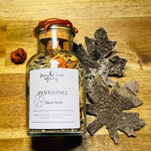 May include: A glass jar with a cork stopper and a white label that reads "Stoney Creek wytchery ABUNDANCE Ritual Herbs Peppermint, Canquefoil, Cinnamon, Clove, Basil, Clove, Ginger Root, Nutmeg". The jar is filled with dried herbs and spices.