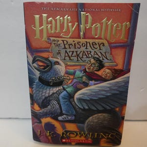 May include: A hardcover book titled Harry Potter and the Prisoner of Azkaban by J.K. Rowling. The cover features a colorful illustration of a person riding a large, winged creature with a beak, set against a warm orange and purple background.