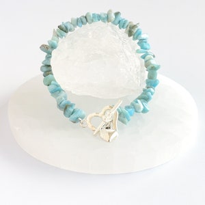May include: A bracelet made of light blue chip stones with a silver heart-shaped clasp. The bracelet features small, irregular-shaped stones in shades of blue and is displayed on a white surface.