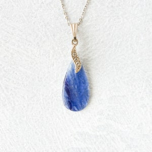 Op de afbeelding: Een zilveren ketting met een druppelvormige blauwe hanger. De hanger is een diepblauwe steen met een goudkleurige decoratieve bovenkant versierd met kleine, heldere steentjes. De ketting hangt aan een fijne zilveren ketting.
