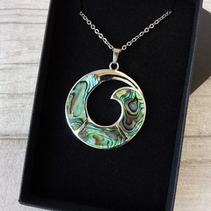 May include: A silver pendant necklace with a circular, wave-shaped design. The pendant is inlaid with iridescent abalone shell pieces in green, blue, and purple hues. The necklace is displayed in a black jewellery box.