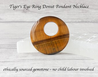 Puede incluir: Collar con colgante de anillo de ojo de tigre. Colgante circular pulido con un agujero en el centro. La piedra preciosa presenta un color marrón cálido con vetas doradas y negras. El texto de la imagen dice "ethically sourced gemstone - no child labour involved."