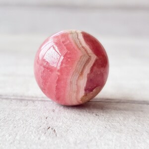 May include: A polished, spherical gemstone with a pink and white banded pattern. The stone has a glossy finish and appears to be a type of mineral. The sphere is set against a light-colored, textured surface.