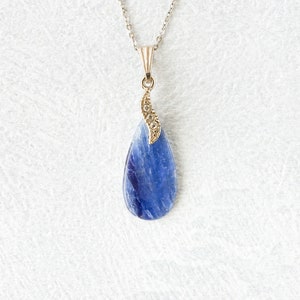 Op de afbeelding: Een zilveren ketting met een druppelvormige hanger. De hanger heeft een levendige blauwe steen met een goudkleurige decoratieve bovenkant. De ketting wordt gepresenteerd op een wit, getextureerd oppervlak.