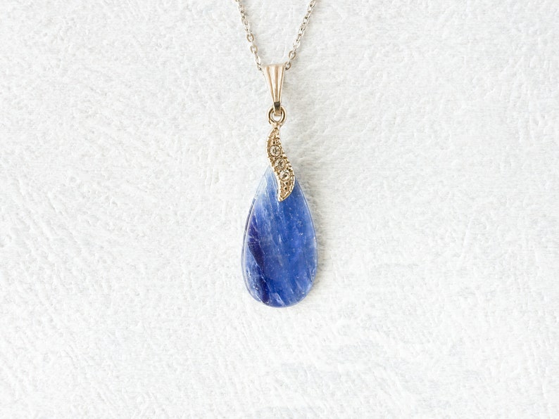 Op de afbeelding: Een zilveren ketting met een druppelvormige hanger. De hanger heeft een levendige blauwe steen met een goudkleurige decoratieve bovenkant. De ketting wordt gepresenteerd op een wit, getextureerd oppervlak.