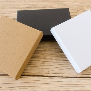 Puede incluir: Tres cajas cuadradas de diferentes colores est&aacute;n dispuestas sobre una superficie de madera marr&oacute;n claro. Las cajas son de color marr&oacute;n natural, negro y blanco. Las cajas est&aacute;n cerradas y parecen vac&iacute;as.