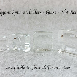 May include: Four clear glass sphere holders in different sizes. Each holder is square with a curved opening. Text reads "Elegant Sphere Holders - Glass - Not Acryl" and "available in four different sizes."