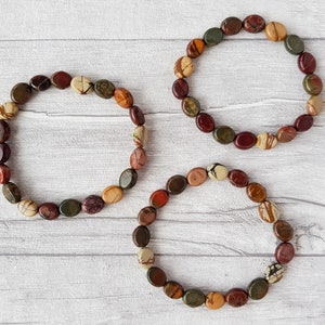 Puede incluir: Tres pulseras hechas de piedras ovaladas pulidas en tonos marr&oacute;n, rojo, verde y crema. Las piedras est&aacute;n ensartadas para formar una forma circular. Las pulseras se exhiben sobre una superficie clara con vetas de madera.