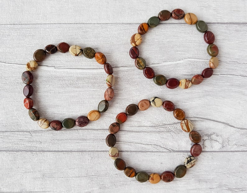 Puede incluir: Tres pulseras hechas de piedras ovaladas pulidas en tonos marr&oacute;n, rojo, verde y crema. Las piedras est&aacute;n ensartadas para formar una forma circular. Las pulseras se exhiben sobre una superficie clara con vetas de madera.