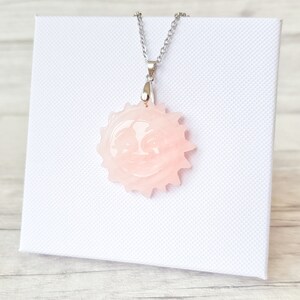 May include: A pink sun pendant necklace with a smiling face design. The pendant is attached to a silver chain. The necklace is displayed on a white textured surface.