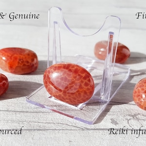 May include: Polished Fire Agate gemstones in shades of red and orange, displayed on a clear acrylic stand. The stones have a glossy finish and a unique, textured pattern. Text on the image reads "Natural & Genuine", "ethically sourced", and "Reiki infused".