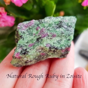 May include: A natural rough ruby in zoisite stone. The stone displays a mix of green, black, and red colours. The text "Natural Rough Ruby in Zoisite" is written in a cursive font at the bottom of the image.