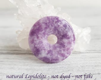 May include: A circular purple and white lepidolite stone pendant with a center hole, positioned in front of a clear quartz crystal cluster. The text below reads: natural Lepidolite - not dyed - not fake - not reconstituted - lovingly Reiki infused.
