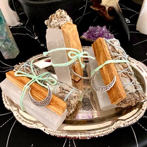 May include: Three bundles of sage smudge sticks with white selenite crystals and silver crescent moon charms on a silver tray. The sage bundles are tied with light blue ribbon.