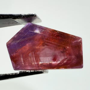 Könnte beinhalten: Ein facettierter Edelstein in Trapezform mit einem Farbverlauf von tiefem Violett bis zu rötlichem Orange. Der Stein weist interne Einschlüsse sowie scharfe, kantige Formen auf und wird von einer Metallpinzette vor einem hellen Hintergrund gehalten.