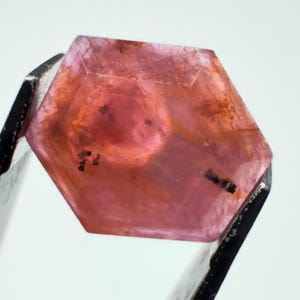 May include: A hexagonal, translucent pink gemstone held by metal tweezers. The stone features a faceted surface with visible internal inclusions, including small dark spots and linear markings within the crystalline structure of the pink gem.