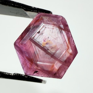 May include: A faceted, pentagonal-shaped pink gemstone held by black metal tweezers. The stone features a translucent appearance with visible internal inclusions, geometric facet lines, and subtle variations in color ranging from deep magenta to pale pink.