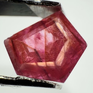 May include: A faceted, pentagonal-shaped gemstone with a deep magenta and reddish-pink hue. The stone features intricate internal geometric patterns and fine linear inclusions visible through its transparent, polished surface.