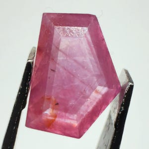 Pode incluir: Uma pedra preciosa rosa lapidada em forma de escudo, segurada por uma pinça de metal. A pedra apresenta um tom magenta translúcido e vibrante com inclusões internas visíveis e estrias naturais por toda a sua estrutura polida.