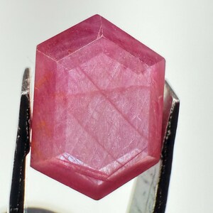 May include: A faceted, hexagonal-shaped pink gemstone held by metal tweezers. The stone displays a translucent, deep rose-pink hue with visible internal inclusions and geometric facets that catch the light across its flat, polished surface.