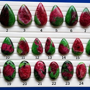 Puede incluir: Una colección de veinticinco piedras preciosas de zoisita pulida con inclusiones de rubí, dispuestas en tres filas sobre una bandeja blanca. Cada piedra presenta un patrón único de colores verde vibrante y rojo intenso, numeradas del 1 al 25.
