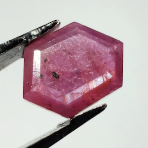May include: A faceted, hexagonal-shaped ruby gemstone with a deep pinkish-red hue and visible internal inclusions. The gemstone is held by a pair of metal tweezers, showcasing its geometric cut, polished surfaces, and natural color variations.