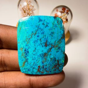 Puede incluir: Un cabujón de piedra preciosa turquesa rectangular que presenta un color base azul cian vibrante con intrincados patrones de vetas de matriz de color marrón oscuro y azul profundo en toda la superficie pulida y lisa.