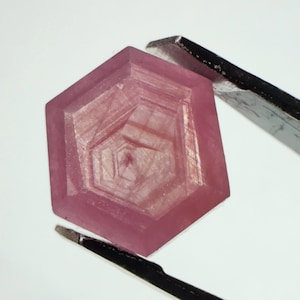 Könnte beinhalten: Ein sechseckiger rosa Edelstein mit facettierter Oberfläche und konzentrischen geometrischen Mustern. Der Stein wird von einer dunklen Metallpinzette gehalten und zeigt ein durchscheinendes Aussehen mit subtilen Lichtreflexen.