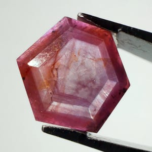 May include: A faceted, pentagonal-shaped gemstone with a deep pink and reddish hue. The stone features internal inclusions and a translucent quality, with visible geometric facets across its surface, held by dark metal tweezers.