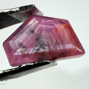 Peut inclure: Une pierre précieuse facettée de forme trapézoïdale avec une teinte rose et rougeâtre translucide. La pierre présente des zones géométriques internes, des inclusions visibles et une surface polie, tenue par une pince métallique.