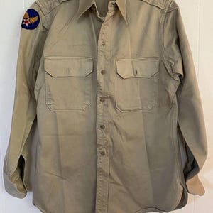 Könnte beinhalten: Khakifarbene Hemd im Militärstil mit zwei Brusttaschen und Knopfverschlüssen. Es hat Schulterklappen und einen Aufnäher auf der linken Schulter mit einem blauen, roten und goldenen Emblem. Das langärmlige Hemd hängt an einem weißen Kleiderbügel.