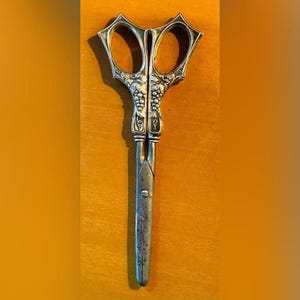 May include: Antique silver scissors with ornate handles. The handles feature a grape vine design. The scissors are made of metal and have a vintage appearance. The blades are straight and the overall design is symmetrical.