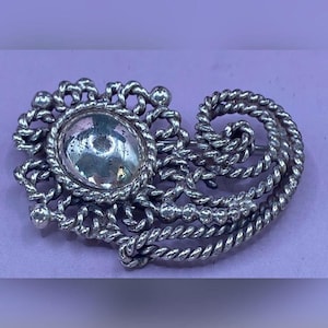 May include: Silver-toned brooch with an oval, clear center stone. The brooch features a detailed, rope-like design around the stone and throughout the piece. The brooch is set against a lavender background.