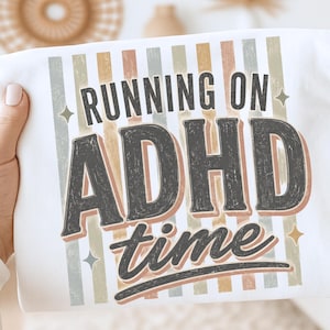 May include: A white t-shirt featuring a graphic design with vertical stripes in muted tones of sage green, tan, and dusty rose. The text reads "RUNNING ON ADHD time" in a dark charcoal, distressed font, accented by small four-pointed star icons.