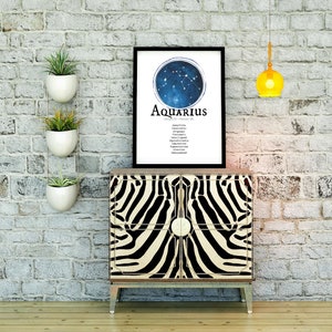 May include: A black framed print with a blue and white illustration of the Aquarius constellation. The print is on a white background and includes the text "Aquarius" and the dates "January 20 - February 18". The print is hanging on a white brick wall above a zebra-print cabinet.