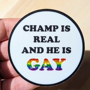 May include: A white round sticker with a black border. The sticker has black text that reads "Champ is real and he is" with the word "gay" in rainbow glitter letters.