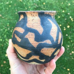 Può includere: Un piccolo vaso in ceramica rotondo, tornito a mano, caratterizzato da un corpo in argilla color cuoio maculato, decorato con spesse linee nere fluide che avvolgono l'esterno. Il vaso presenta un bordo ampio e leggermente svasato.