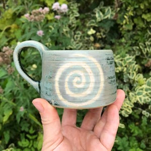 May include: A light blue ceramic mug with a prominent white spiral design on the side. The mug features a textured, speckled finish with darker blue accents near the handle and base, showcasing a cylindrical shape and a curved side handle.