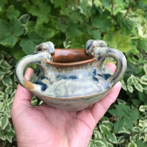 May include: A small, round ceramic bowl with two arched handles. The exterior features a mottled glaze in shades of light blue, grey, and tan, while the interior rim is finished in a smooth, solid brown glaze. The bowl has a wide, open top.