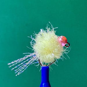 May include: A fishing fly lure featuring a fluffy, pale yellow synthetic body with iridescent, translucent fibers extending from the back. A small, shiny, metallic pink bead is attached near the eye of the dark metal hook, which is mounted on a blue tool.