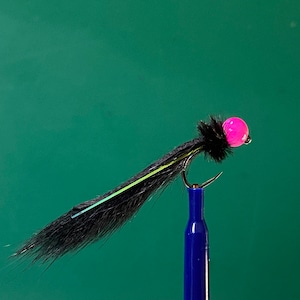 Puede incluir: Primer plano de una mosca de pesca con cuerpo negro, cabeza de cuentas rosa y anzuelo curvo. La mosca tiene un cuerpo negro y plumoso con un fino acento amarillo y azul. El anzuelo está sujeto por una herramienta azul sobre un fondo verde.